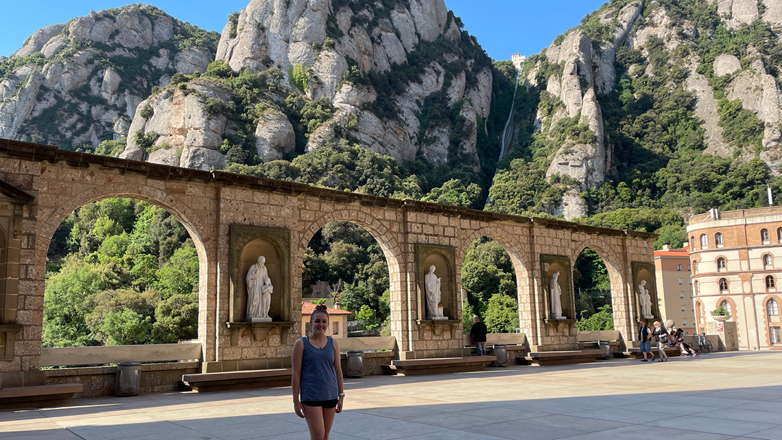 Student stands in front of ornate building with verdant mountains behind in Montserrat, Catalonia